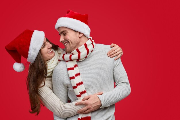 Joyful couple in red christmas hat and scarf hugging and smiling