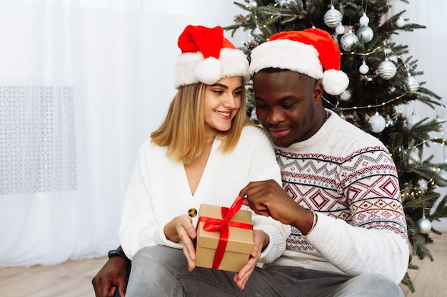 Joyful cozy moments in winter holidays. A multinational couple opens a Christmas present. A loving young black man and a white woman are sitting next to the Christmas tree