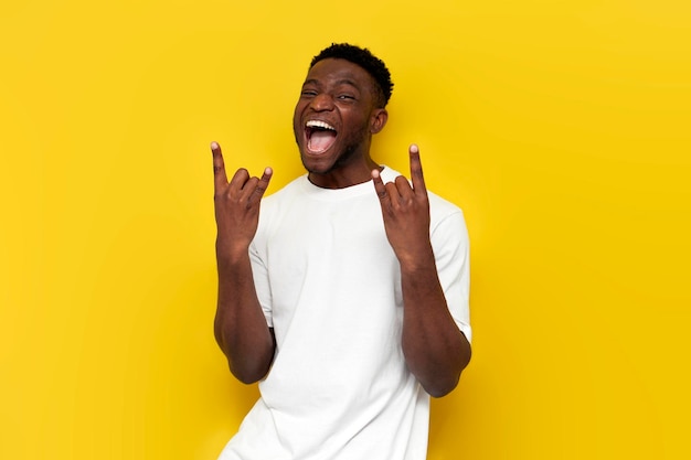 Joyful crazy african american man shows rock gesture with his hands and shouts on yellow background