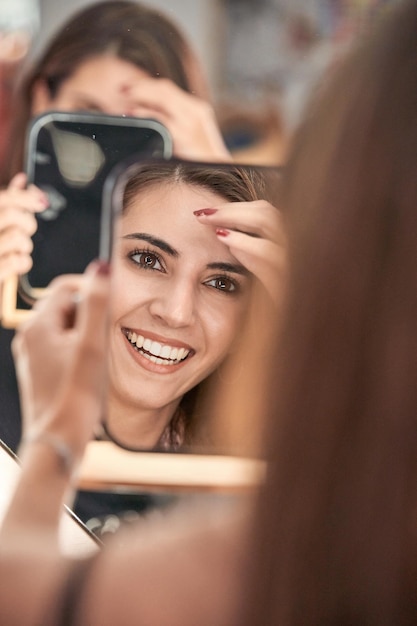 Photo joyful crop female with long hair looking at reflection in mirror with smile while sitting in modern light mirror during appointment