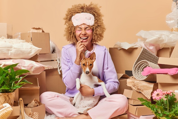 Joyful curly haired woman sits in lotus pose with pet laughs\
out happily applies hydrogel patches under eyes surrounded by\
cardboard boxes full of belongings moves to new apartment real\
estate