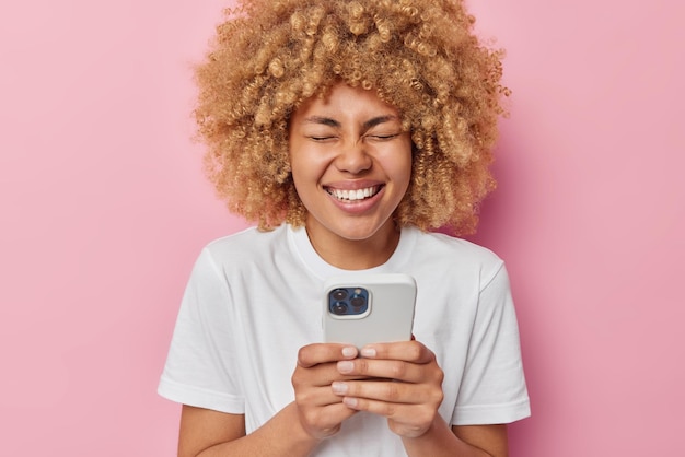 Joyful curly haired woman types text messages surfs internet and laughs sincerely reads funny content in internet giggles positively dressed in white casual t shirt isolated over pink background