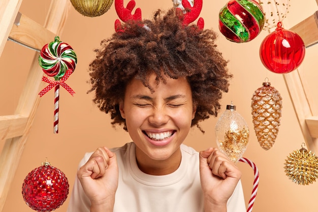 Photo joyful curly haired young woman closes eyes and clenches fists celebrates success wears red reindeer and casual t shirt poses against beige background with new year toys. christmas time concept