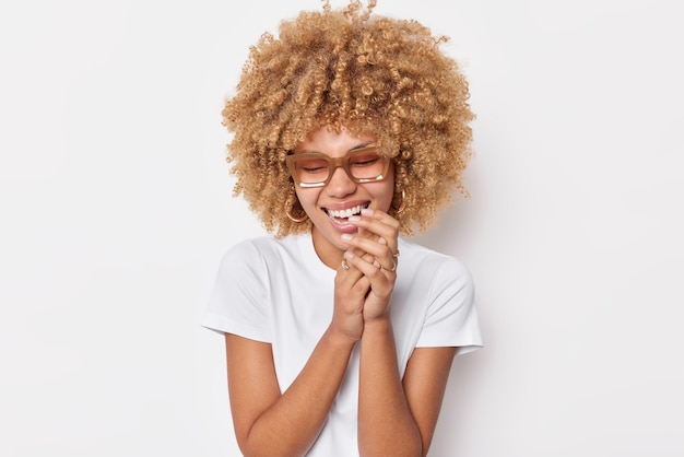 Joyful curly haired young woman laughs happily keeps hands near face feels very happy smiles broadly wears casual t shirt and spectacles isolated over white background hears something very funny