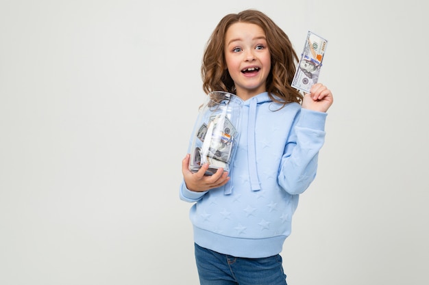 Joyful cute girl with a glass jar and money on a white wall with blank space.