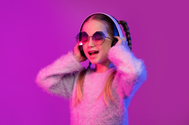 Joyful cute little girl using wireless headphones in neon light