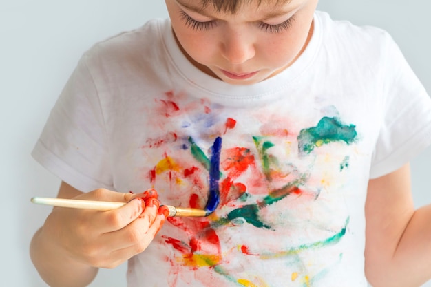 カラフルな塗装の手で5〜6歳のうれしそうなかわいい未就学児の小さな男の子。子供が指で描いたTシャツ。絵を描くこと、教育すること、そして子どもの発達。幸せな子供時代