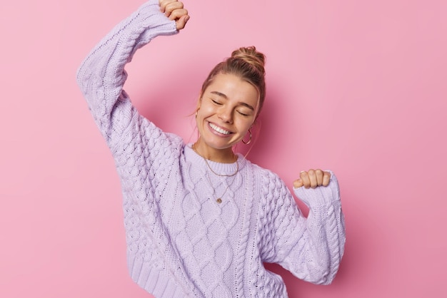 Joyful dancing woman shakes arms has eyes closed expresses happiness and upbeat emmotions wears loose knitted sweater isolated over pink background feels carefree dances with rhythm of music