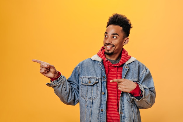 Joyful dark-skinned guy in denim jacket points his finger to side