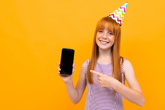 Joyful european red-haired girl shows the phone screen over yellow 