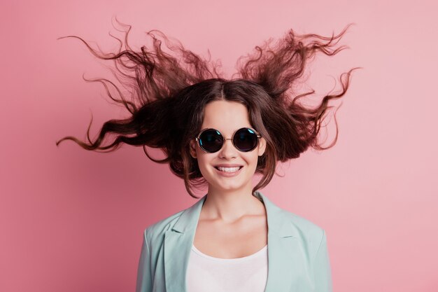 Joyful excited surprised young woman with flying hair wear blazer sunglass on pink wall