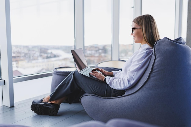 Gioiosa donna dai capelli biondi in camicia bianca che digita il testo della pubblicazione utilizzando un computer portatile che lavora in remoto ragazza allegra con gli occhiali condivide contenuti positivi e file multimediali sul netbook