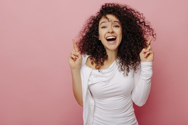 Foto gioiosa giovane ragazza dalla pelle chiara incrocia le dita con entrambe le mani sorridendo ampiamente su sfondo rosa bruna dai capelli gonfi in camicetta bianca emozioni di bellezza concetto di persone