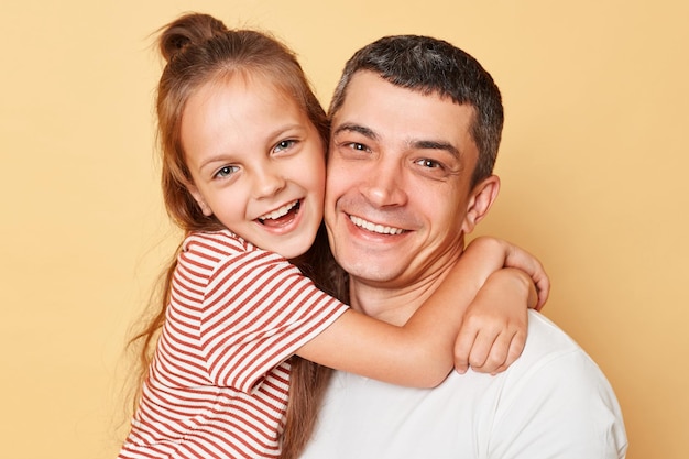 Joyful family father and daughter wearing casual tshirts standing hugging each other looking at camera with happy faces isolated over beige background