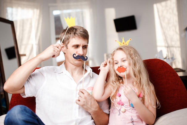 Joyful father and daughter with paper crowns and mustaches while sitting togheter on red chair at home.