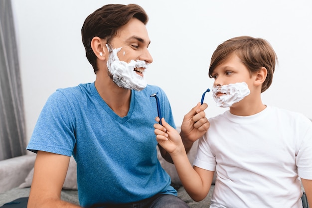 Joyful father and son with foam on faces shaves each other.