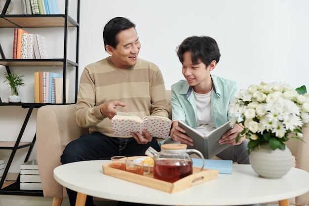 Joyful father and teenage son reading and discussing books with interesting novels at home when drinking tea