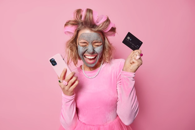 Joyful female model has fun holds credit card while paying with
mobile phone application applies hair rollers and beauty clay mask
for skin care purchases something wears dress and necklace