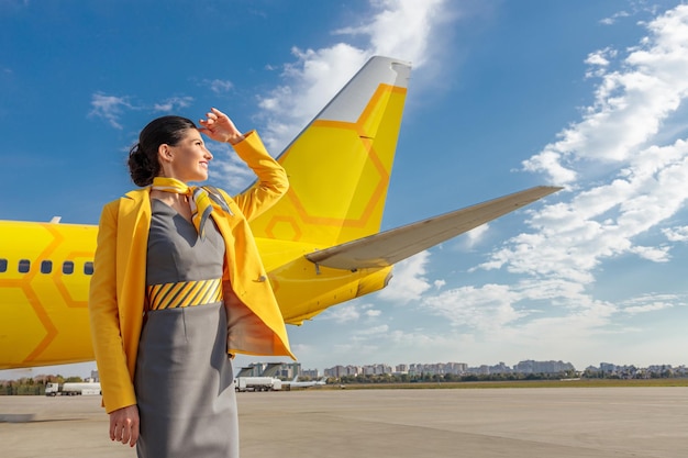 彼女の手で太陽から目を覆い、曇り空に立って笑っている航空会社の制服を着た楽しい客室乗務員