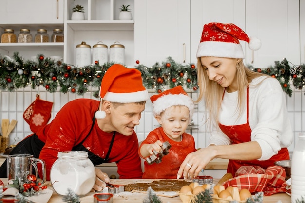 Joyful friendly family wearing Christmas hats are cooking gingerbread in the kitchen Excited little boy in santa hat is going to cut dough with cookie cutter