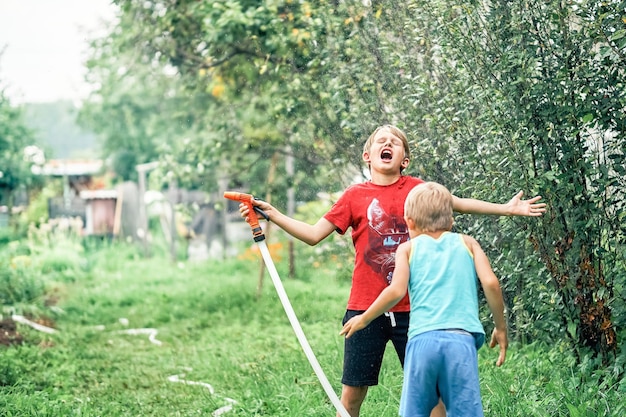 Joyful funny boy with cute little brother plays with water hose in green garden in summerhouse yard