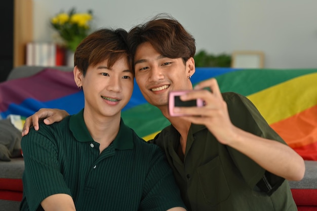 Joyful gay couple taking a selfie with compact camera in living room rainbow flag in background LGBT love and human rights concept