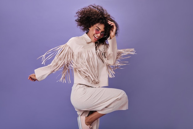 Joyful girl in beige suit and glasses jumping on purple background Cool woman in jacket and pants dancing on isolated backdrop