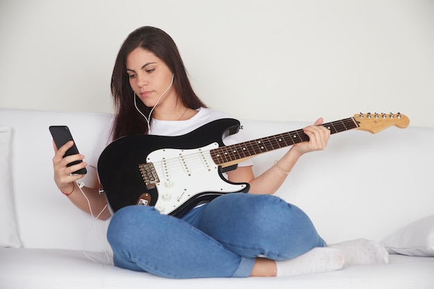 Joyful girl in earphones watching electric guitar tutorial on\
mobile phone