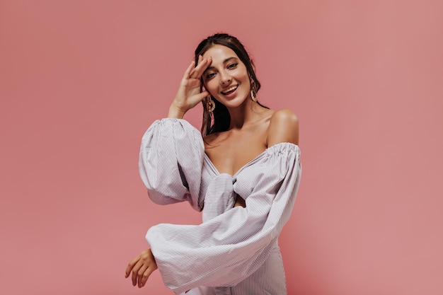 Joyful girl in good mood with black wavy hair in gold cool earrings and lilac striped clothes looking into camera and smiling