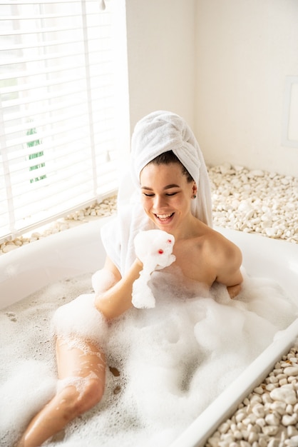 Joyful girl is playing with foam while taking a bath at spa. She lies in the bathroom and laughs