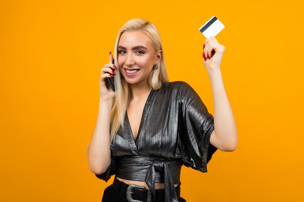 joyful girl makes purchases by phone holding a credit card with a mockup on yellow wall