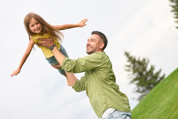 joyful girl playing with her daddy in the green park stretching out her hands pretending flying