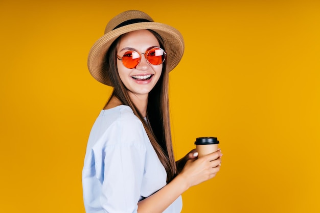 La ragazza allegra in occhiali da sole tiene il caffè nelle sue mani. sfondo giallo
