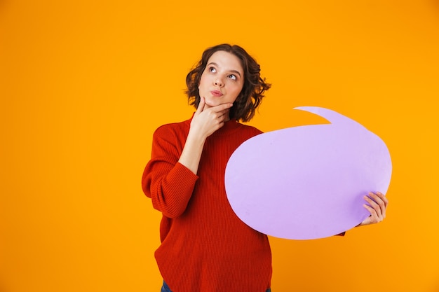 Joyful girl wearing sweater holding thought bubble with copyspace while standing isolated on yellow