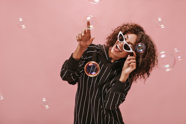 Joyful girl with curly brunette hairstyle in long sleeve black dress and white cool sunglasses smiling and posing with bubble on isolated wall..