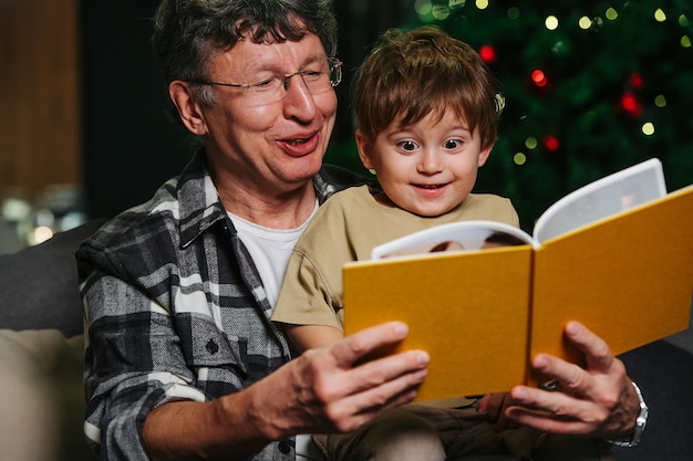 Joyful grandpa and surprised little boy sitting on a sofa looking through a photo album. Christmas tree in background.