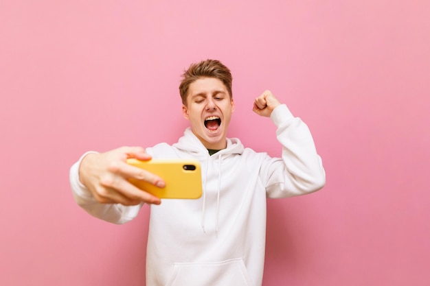 Joyful guy gamer stands with a smartphone in his hand on a pink background