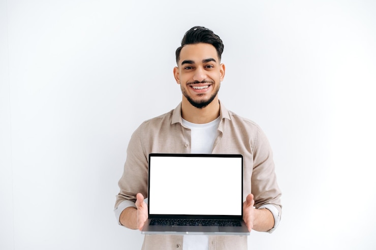 Photo joyful handsome indian or arabian young man student or freelancer stands on white isolated background holding open laptop with blank white mockup screen for presentation looking at camera smiling