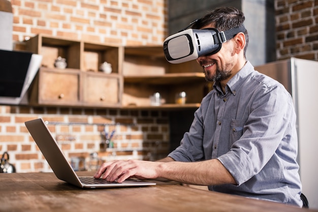 Радостный красавец, использующий свой ноутбук во время ношения устройства VR