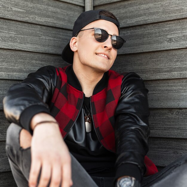 Joyful handsome young hipster man in torn gray jeans in a black cap in a trendy red and black checkered jacket in stylish sunglasses sits near a vintage wooden wall and enjoys the weekend