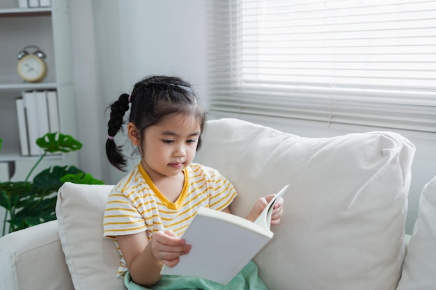 Joyful happy asian child baby girl smiling and reading book while sitting on couch sofa in living room at home girl relex reading book smile at sofa in the house back to school concept