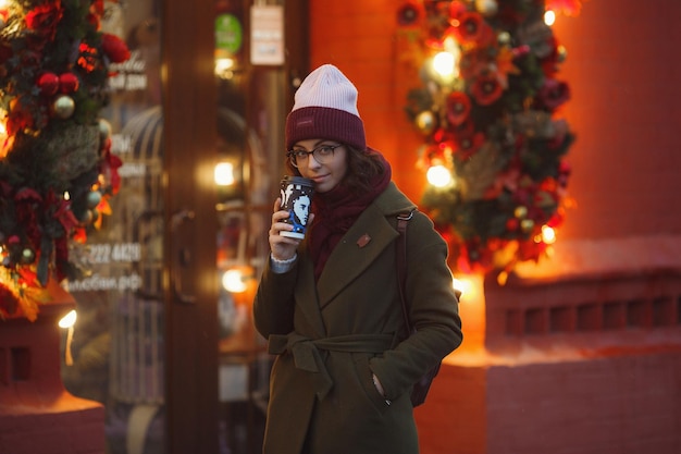 사진 거리에서 눈이 내리는 날씨를 즐기는 즐거운 행복한 소녀 미소 커피를 마시고 인생을 즐기는 세련된 모피 코트 니트 모자와 넓은 스카프를 착용 축제 분위기 크리스마스 분위기 진정한 행복