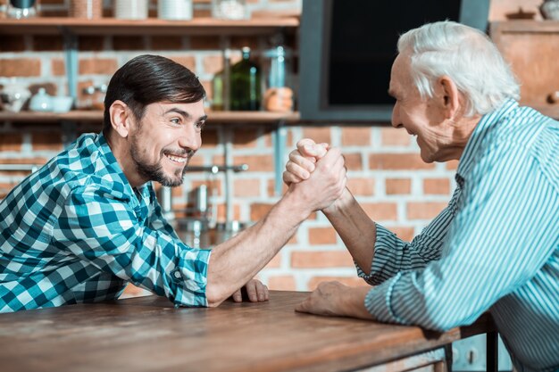 Joyful happy nice father and son holding each others hands and doing arm wrestling while playing together