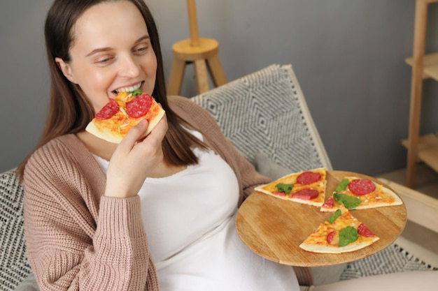 Joyful happy pregnant woman eating pizza alone at home female satisfied with timely food delivery sitting on sofa in living room enjoying fast food during pregnancy