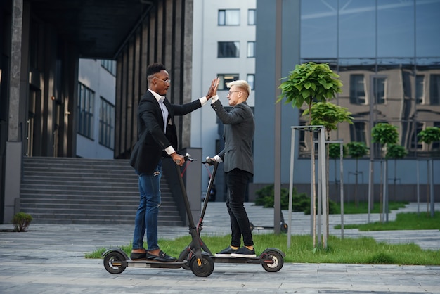 Joyful international office workers riding on electric scooters and give high fives at motion.