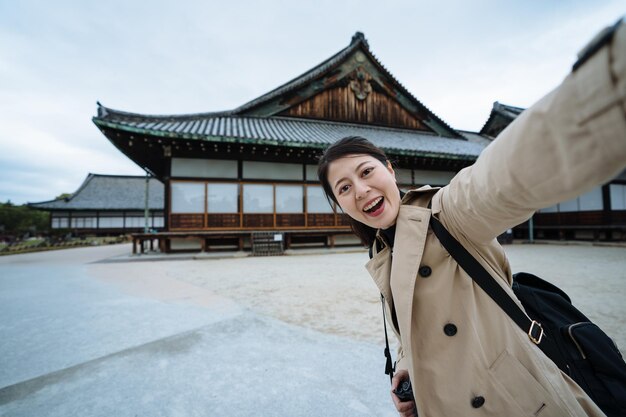 Joyful korean visitor is taking a selfie with traditional
religious building in kyoto. excited female tourist looking at
camera is taking picture to commemorate her trip to nijo
castle.