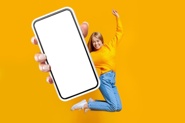 Joyful lady jumping in air on orange background showing smartphone