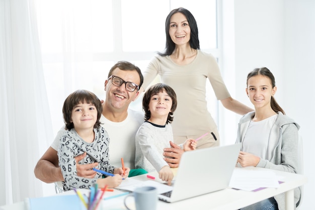 Joyful latin family with children smiling at camera while spending time together at home. Father working from home, using laptop and watching kids. Technology, family concept