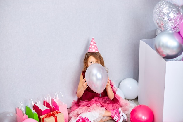 Joyful little birthday girl in pink dress and hat covers his face with a balloon at home party