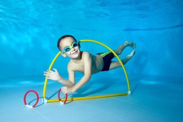 A joyful little boy dives under the water in the pool swims through the hoop and pulls out toys